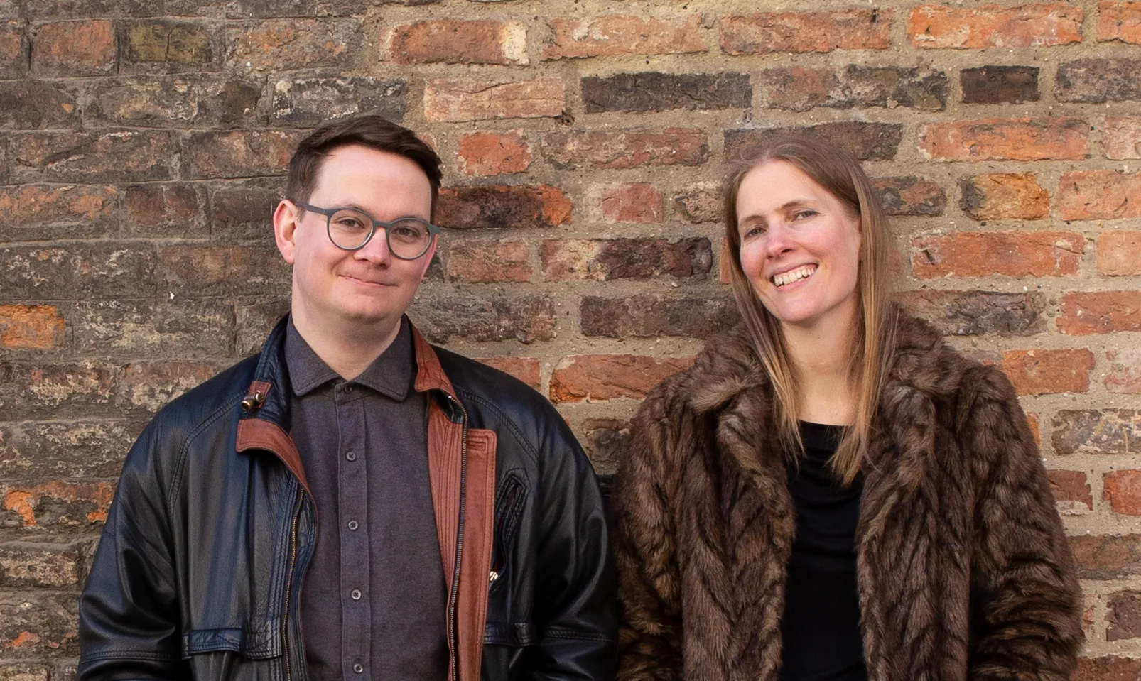 Alex and Rachel leaning against a brick wall, smiling at the camera